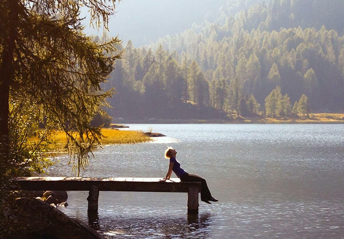 A person sitting on the edge of a wooden dock by a calm lake, surrounded by pine trees and mountains in the background. The world is listening.