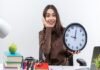 A woman sits at a cluttered desk, smiling while holding a large clock. Books, an apple, and stationery are visible, symbolizing growth. The word "productivity" is at the bottom.