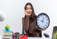 A woman sits at a cluttered desk, smiling while holding a large clock. Books, an apple, and stationery are visible, symbolizing growth. The word "productivity" is at the bottom.