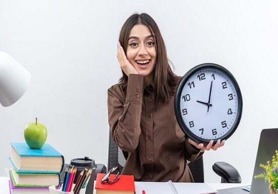 A woman sits at a cluttered desk, smiling while holding a large clock. Books, an apple, and stationery are visible, symbolizing growth. The word "productivity" is at the bottom.