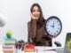 A woman sits at a cluttered desk, smiling while holding a large clock. Books, an apple, and stationery are visible, symbolizing growth. The word "productivity" is at the bottom.