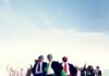 A group of people in business attire, some wearing colorful capes and masks, stand confidently with arms raised against a city skyline, embodying the power of teamwork.