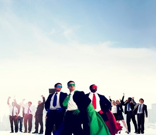 A group of people in business attire, some wearing colorful capes and masks, stand confidently with arms raised against a city skyline, embodying the power of teamwork.