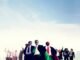 A group of people in business attire, some wearing colorful capes and masks, stand confidently with arms raised against a city skyline, embodying the power of teamwork.