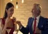 A man in a suit discreetly hands a card to a woman in a red dress at their restaurant table, laden with salads and drinks, hinting at the beginning of an intriguing Sugar Daddy and sugar baby arrangement.