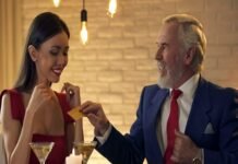 A man in a suit discreetly hands a card to a woman in a red dress at their restaurant table, laden with salads and drinks, hinting at the beginning of an intriguing Sugar Daddy and sugar baby arrangement.