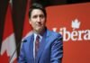 A person in a blue suit and red tie speaks at a podium with a red background displaying the word "Liberal." Trudeau resigns.