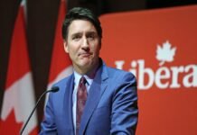 A person in a blue suit and red tie speaks at a podium with a red background displaying the word "Liberal." Trudeau resigns.