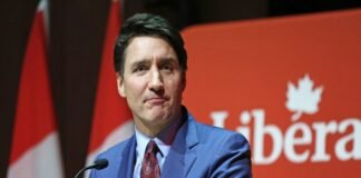 A person in a blue suit and red tie speaks at a podium with a red background displaying the word "Liberal." Trudeau resigns.