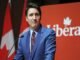 A person in a blue suit and red tie speaks at a podium with a red background displaying the word "Liberal." Trudeau resigns.