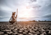 A farmer crouches on cracked, dry ground under a cloudy sky, holding a long stick. Dressed in light clothing and a hat, searching for answers in the parched earth and committing suicide.