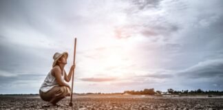 A farmer crouches on cracked, dry ground under a cloudy sky, holding a long stick. Dressed in light clothing and a hat, searching for answers in the parched earth and committing suicide.