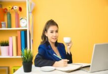 A person sits at a desk, holding a cup and writing in a notebook. A laptop, a plant, shelves with books, and freelance jobs for beginners with no experience