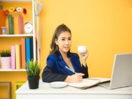 A person sits at a desk, holding a cup and writing in a notebook. A laptop, a plant, shelves with books, and freelance jobs for beginners with no experience