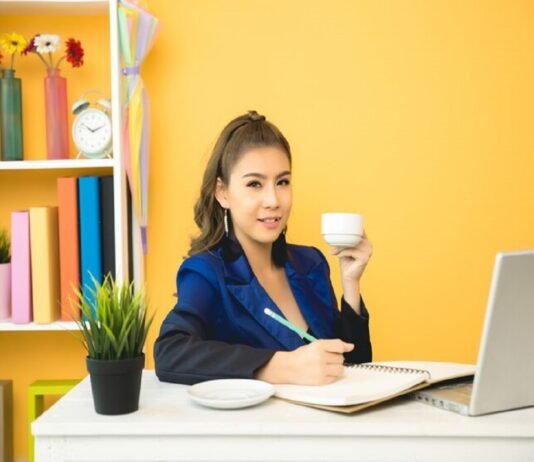 A person sits at a desk, holding a cup and writing in a notebook. A laptop, a plant, shelves with books, and freelance jobs for beginners with no experience