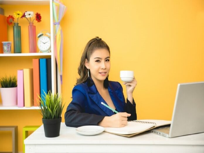 A person sits at a desk, holding a cup and writing in a notebook. A laptop, a plant, shelves with books, and freelance jobs for beginners with no experience