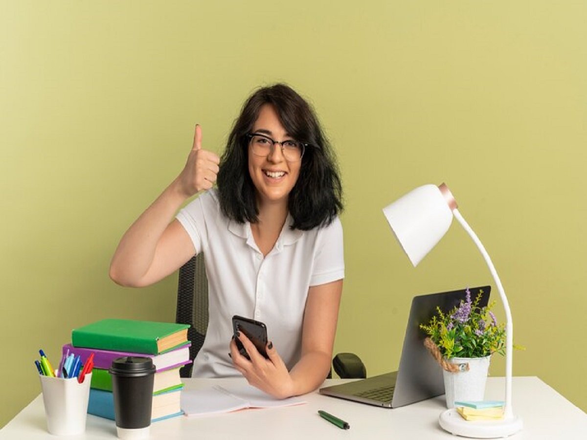 A person sits at a desk with a laptop, books, and a plant, holding a phone and giving a thumbs up for freelance jobs for beginners with no experience.