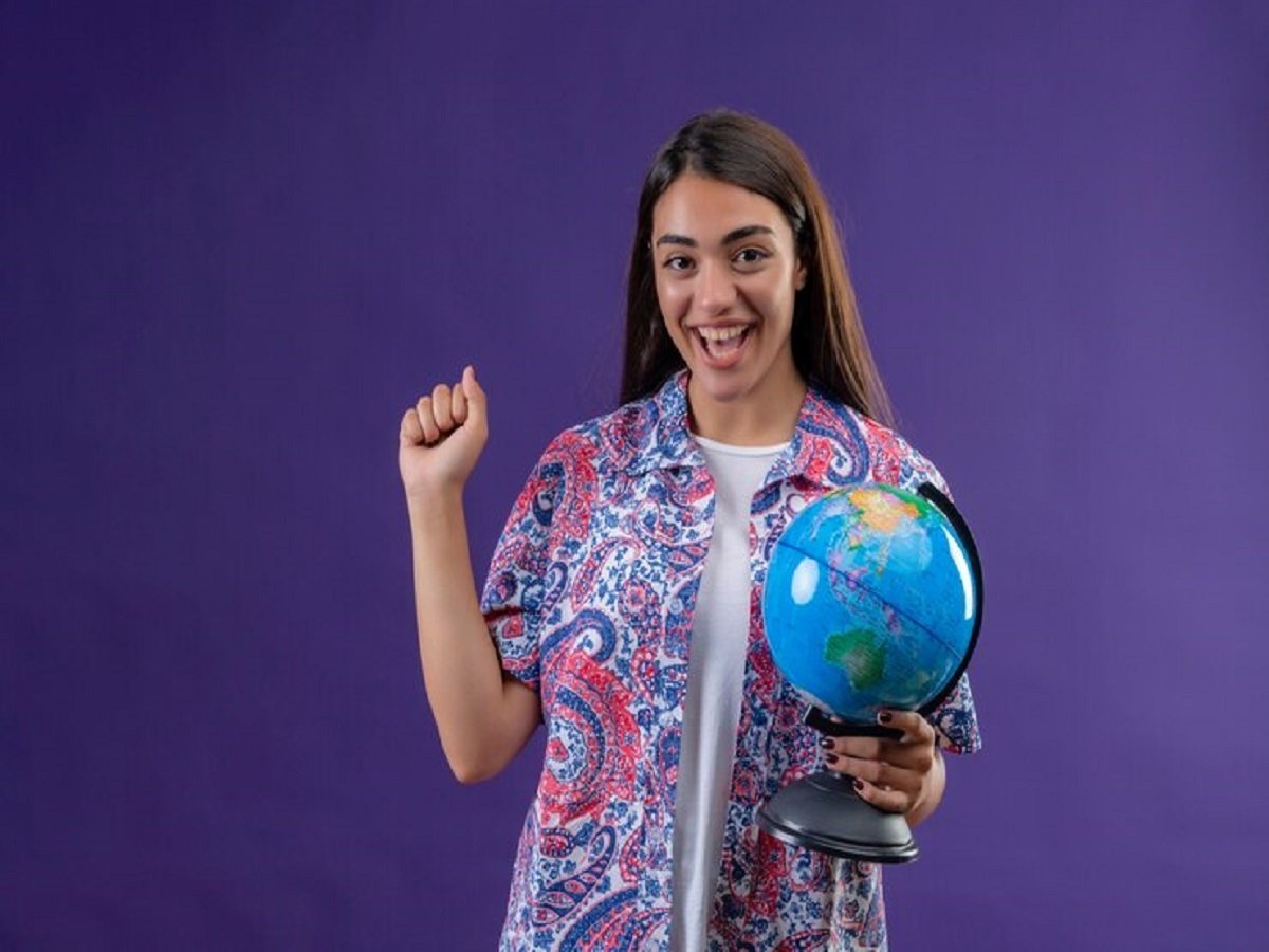 A smiling person, representing Indian immigrants, stands against a purple background, holding a globe in one hand and raising the other. They wear a colorful patterned shirt that highlights their vibrant cultural roots.