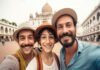 Three Indian immigrants wearing hats smile for a selfie in front of a historic building with domes and arches.