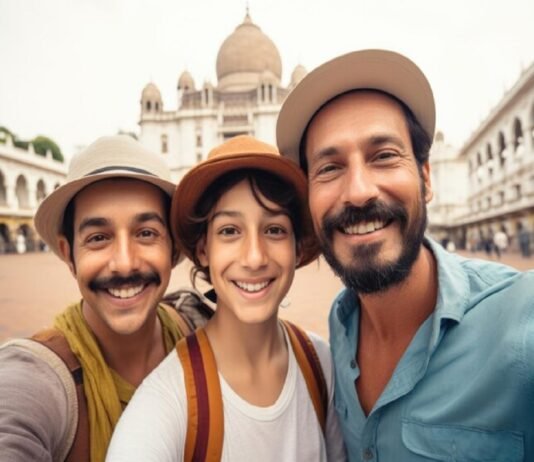 Three Indian immigrants wearing hats smile for a selfie in front of a historic building with domes and arches.