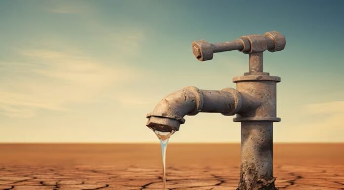 A rusty faucet protrudes from cracked, dry ground, releasing a small stream of water into a shallow puddle under a clear, blue sky—a haunting emblem of water scarcity reminiscent of the Indian water crisis.