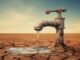 A rusty faucet protrudes from cracked, dry ground, releasing a small stream of water into a shallow puddle under a clear, blue sky—a haunting emblem of water scarcity reminiscent of the Indian water crisis.