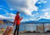 A woman in a red coat looks at a map by a lakeside, pulling a suitcase. As she navigates through the picturesque mountains and clouds under the blue sky, she dreams of finding stability amidst the housing crisis living in Canada.