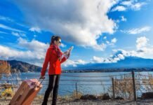 A woman in a red coat looks at a map by a lakeside, pulling a suitcase. As she navigates through the picturesque mountains and clouds under the blue sky, she dreams of finding stability amidst the housing crisis living in Canada.