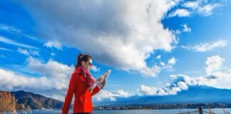 A woman in a red coat looks at a map by a lakeside, pulling a suitcase. As she navigates through the picturesque mountains and clouds under the blue sky, she dreams of finding stability amidst the housing crisis living in Canada.