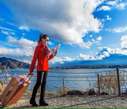 A woman in a red coat looks at a map by a lakeside, pulling a suitcase. As she navigates through the picturesque mountains and clouds under the blue sky, she dreams of finding stability amidst the housing crisis living in Canada.