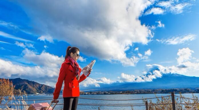 A woman in a red coat looks at a map by a lakeside, pulling a suitcase. As she navigates through the picturesque mountains and clouds under the blue sky, she dreams of finding stability amidst the housing crisis living in Canada.