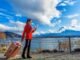 A woman in a red coat looks at a map by a lakeside, pulling a suitcase. As she navigates through the picturesque mountains and clouds under the blue sky, she dreams of finding stability amidst the housing crisis living in Canada.