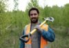 A man in an orange safety vest, reminiscent of migrant workers' attire, holds a shovel over his shoulder as he stands in a grassy area with trees in the background.