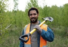 A man in an orange safety vest, reminiscent of migrant workers' attire, holds a shovel over his shoulder as he stands in a grassy area with trees in the background.