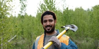 A man in an orange safety vest, reminiscent of migrant workers' attire, holds a shovel over his shoulder as he stands in a grassy area with trees in the background.