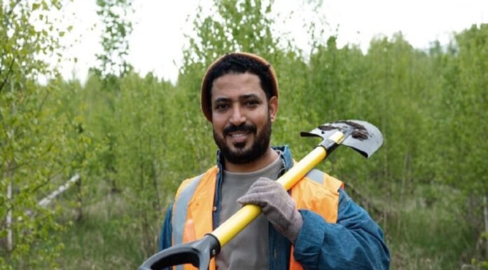 A man in an orange safety vest, reminiscent of migrant workers' attire, holds a shovel over his shoulder as he stands in a grassy area with trees in the background.