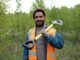 A man in an orange safety vest, reminiscent of migrant workers' attire, holds a shovel over his shoulder as he stands in a grassy area with trees in the background.