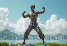 A bronze statue of a martial artist strikes a dynamic fighting pose, set against a waterfront cityscape where other statues stand sentinel under cloudy skies and distant mountains.