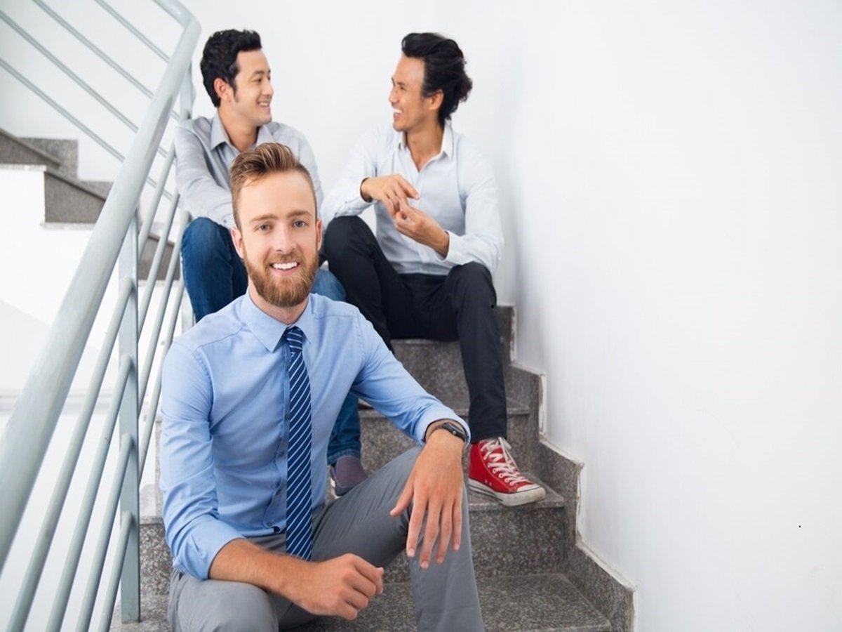 Three unskilled men sitting on stairs, two chatting merrily in the background, while one smiles confidently at the camera.