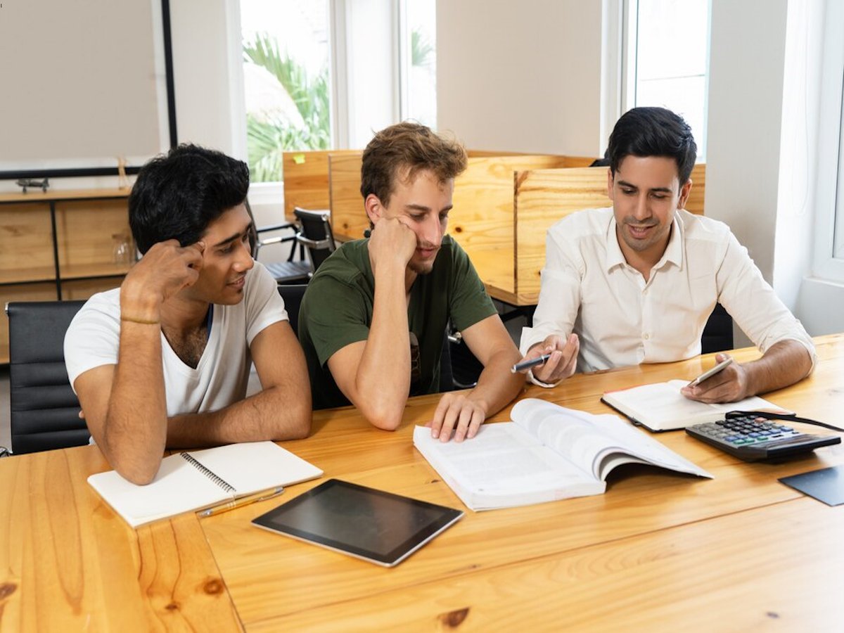 Three underemployed men sit at a wooden table in a bright room, unskilled, discussing documents and using tablets and a calculator.